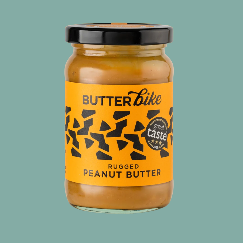 A glass jar of Butterbike Rugged Peanut Butter with a bright yellow label and black lid, centered on a solid teal background.