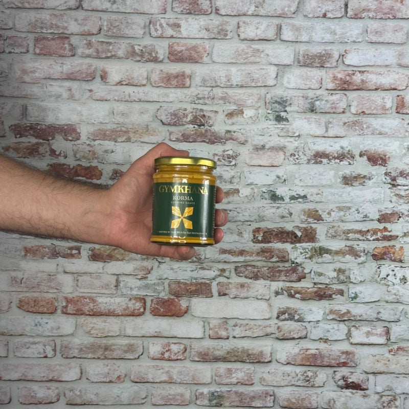 Hand holding a Gymkhana - Korma Sauce jar with gold lid against a brick wall