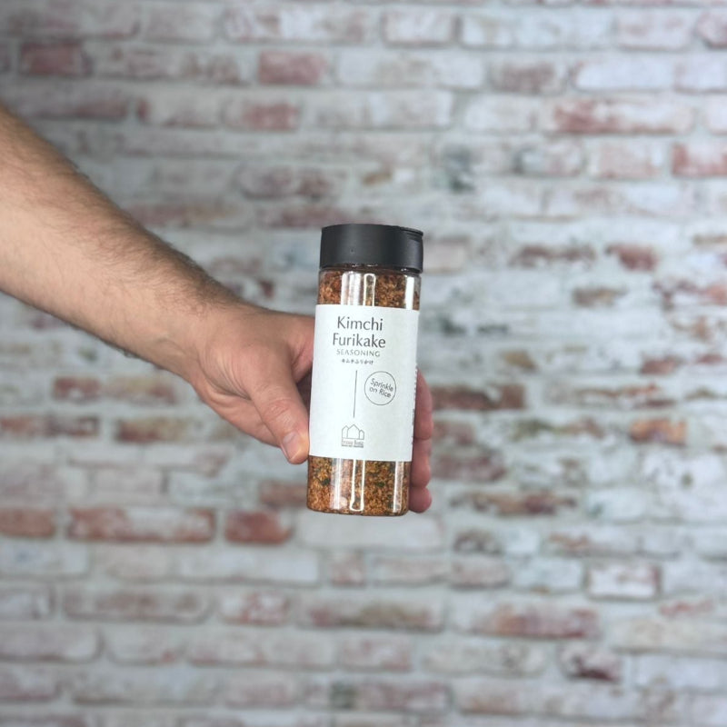 Hand holding a jar labeled 'Kimchi Furikake' against a brick wall.