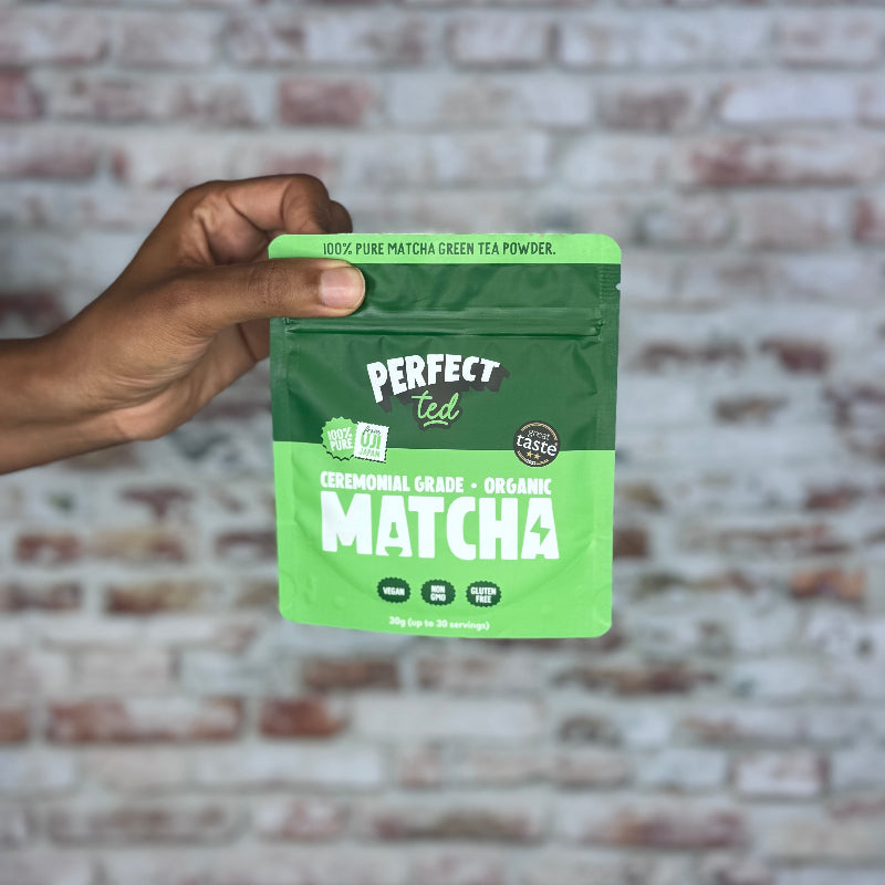Hand holding a green package of Perfect Tea Matcha against a brick wall.