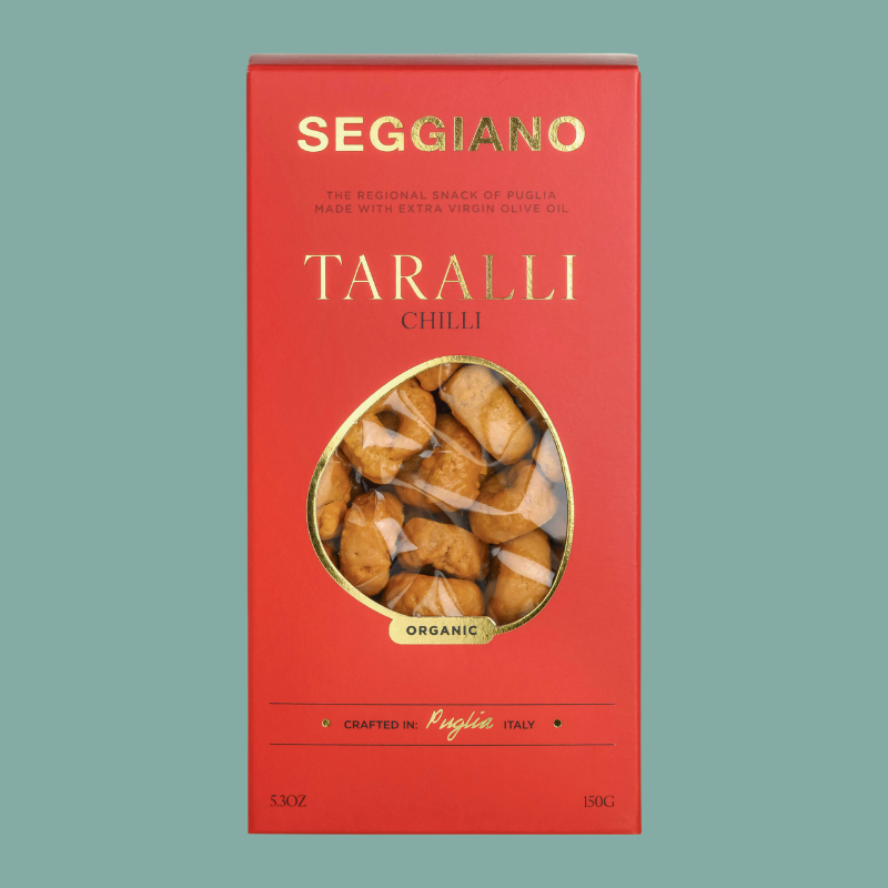 A red box of Seggiano Taralli Chilli, featuring gold lettering and a circular clear window showing the dried chili peppers inside, set against a solid teal background.