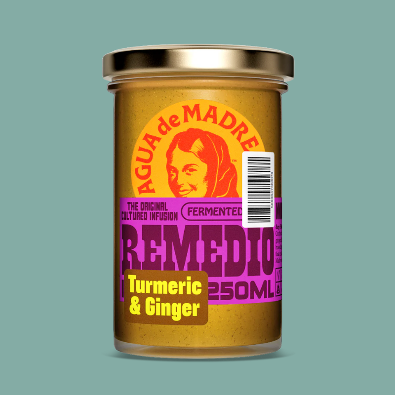 A 250ml glass jar of Agua de Madre Remedio Turmeric & Ginger on a solid teal background. The jar has a gold lid and a colorful label featuring a stylized illustration of a woman's face and bold text in orange, purple, and yellow.