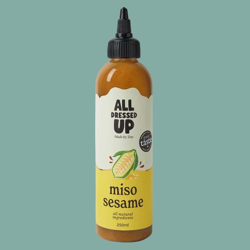 A 250ml squeeze bottle of All Dressed Up Miso Sesame dressing, featuring a black cap and a label with a white top half and a yellow bottom half, set against a solid teal background.
