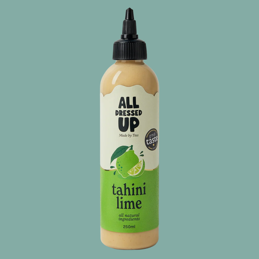 A bottle of All Dressed Up Tahini Lime dressing on a solid teal background. The bottle contains a light tan sauce, has a cream and green label with a lime illustration, and a black squeeze cap.