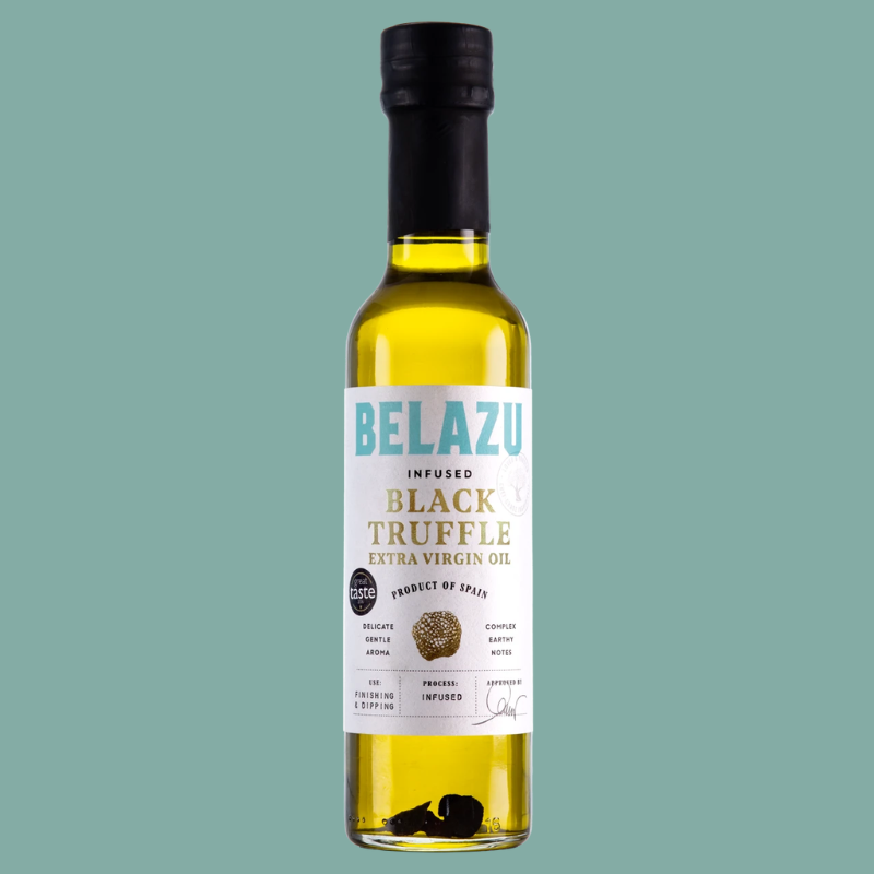 A bottle of Belazu Infused Black Truffle Extra Virgin Oil against a solid sage green background. The bottle is filled with golden-yellow oil and features a white label with blue and gold text, sealed with a black cap.