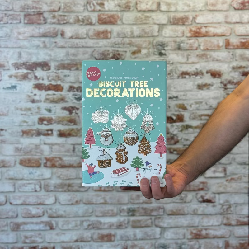 Hand holding a book titled 'Biscuit Tree Decorations' against a brick wall.