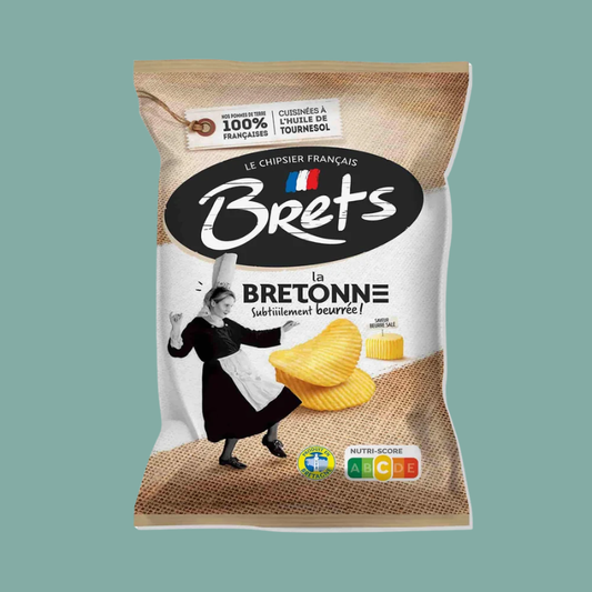 A bag of Brets 'la Bretonne' potato chips on a solid teal-green background. The packaging features a rustic burlap-textured design, a black oval logo with white 'Brets' lettering, and an illustration of a woman in a black dress and white apron dancing excitedly next to yellow potato chips.