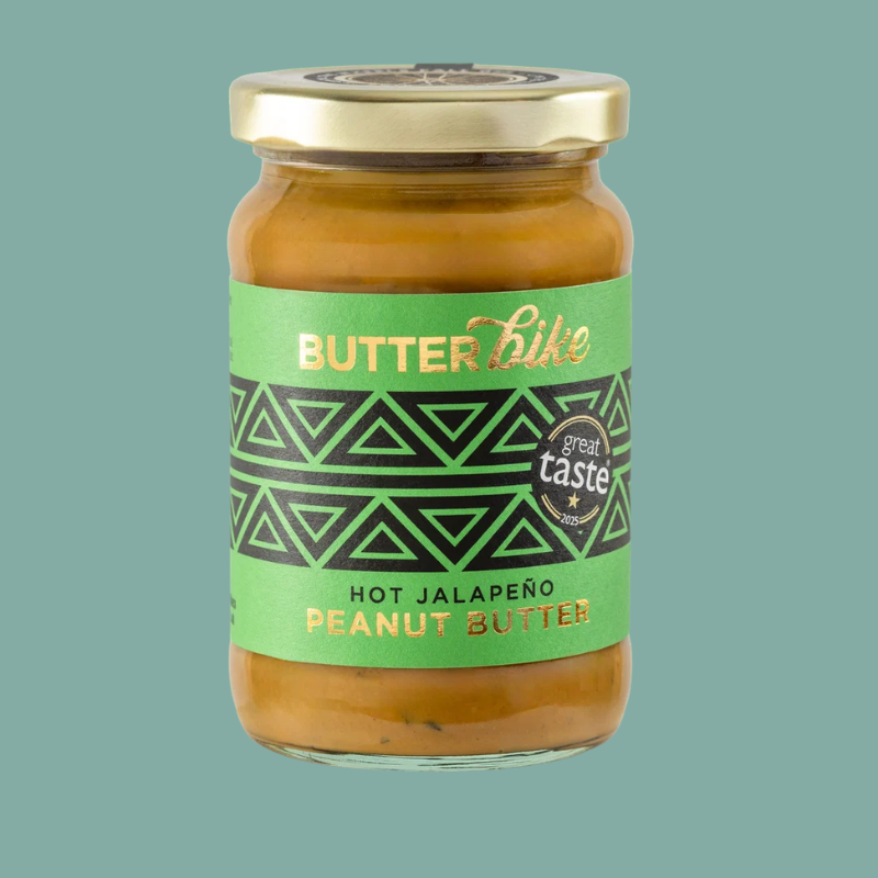 A glass jar of Butterbike Hot Jalapeño Peanut Butter with a green and black label and a gold lid, set against a solid teal background.