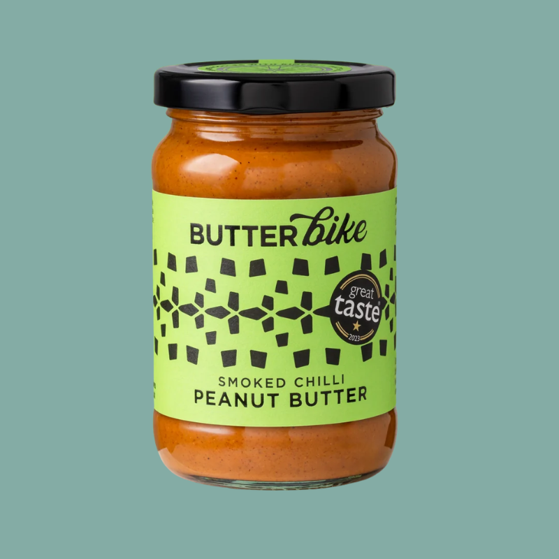 A jar of Butterbike Smoked Chilli Peanut Butter on a solid teal background. The product is in a clear glass jar showing the orange-brown peanut butter inside, with a bright lime green label and a black lid.