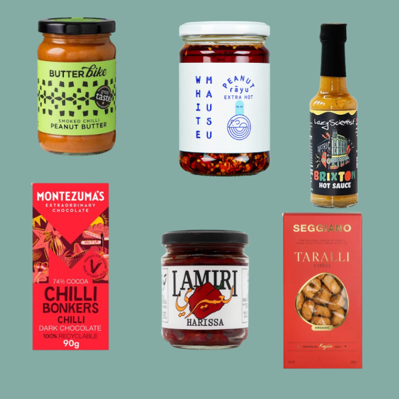 A collage of six packaged food products arranged on a flat muted green background: a jar of Butterbike Peanut Butter, a jar of Whitr Mausu Extra Hot Sauce, a bottle of Brixton Hot Sauce, a Montezuma's Chilli Bonkers chocolate bar, a jar of Lamri Harissa, and a red box of Seigiano chilli Taralli.