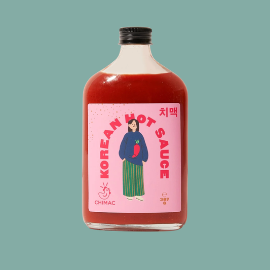 A bottle of Chimac Korean Hot Sauce with a pink label and black cap, filled with red sauce, centered on a solid teal-green background.