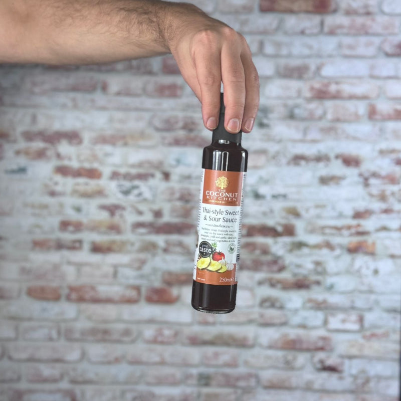 Hand holding a bottle of Coconut Kitchen Thai Sweet & Sour Sauce against a brick wall background