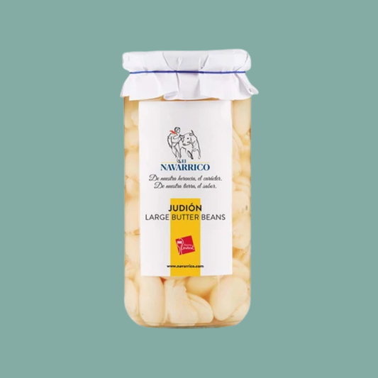 A glass jar of Judún Large Butter Beans from the brand 'Suelo Navarrico', featuring a white paper label and a white fabric-wrapped lid, set against a solid teal background.