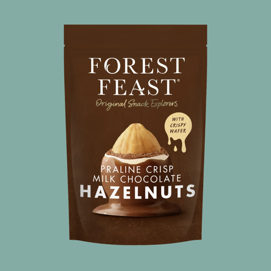 Forest Feast Praline Crisp Milk Chocolate Hazelnuts in a brown stand-up pouch with white text and a chocolate-hazelnut illustration, shown on a muted teal background.