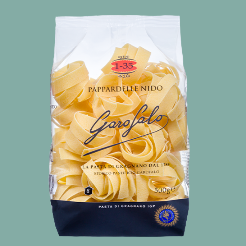 A 500g bag of Garofalo Pappardelle Nido pasta, showing the yellow flatpasta through a clear window in a white and navy blue package, set against a solid sage green background.