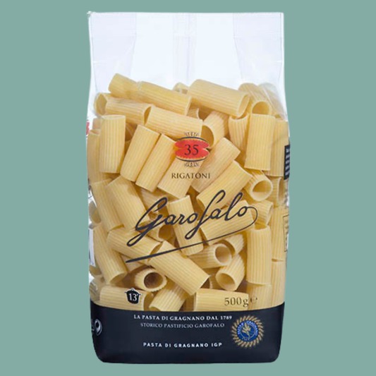 A 500g bag of Garofalo Rigatoni pasta, showing the yellow pasta pieces inside a clear bag with a black and white label, set against a solid sage green background.