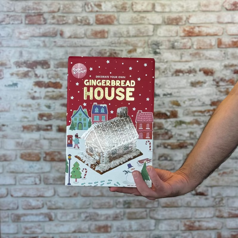 Hand holding a gingerbread house-themed product packaging in front of a brick wall.