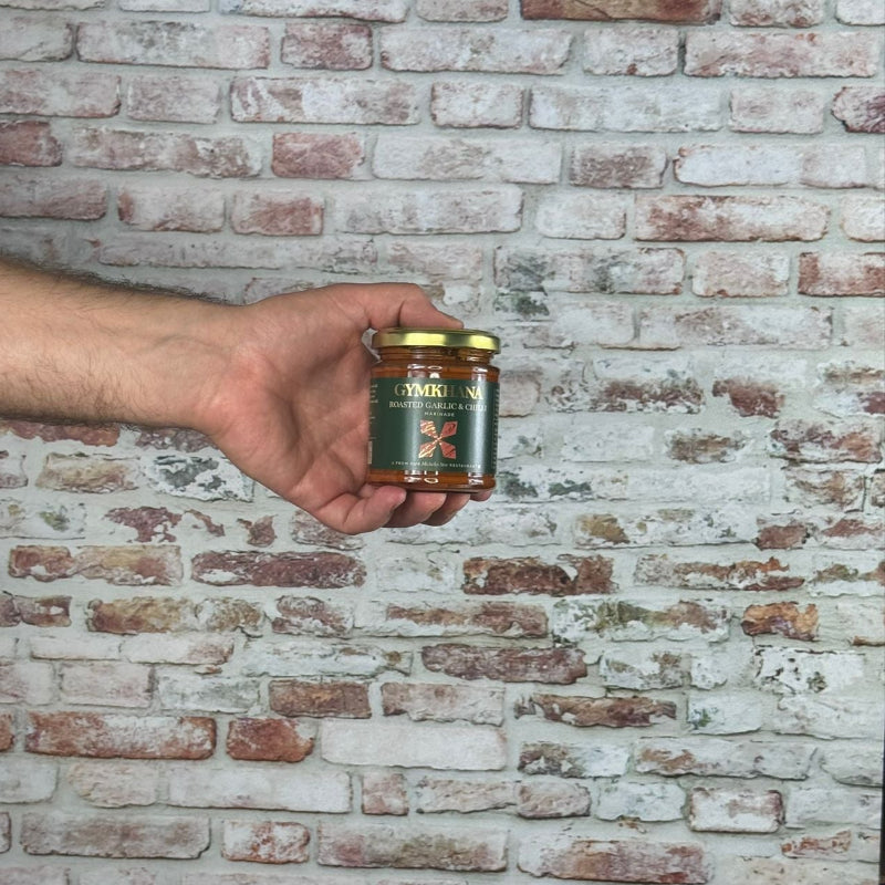Hand holding a jar of Gymkhana - Roast Chilli Garlic Marinade in front of a brick wall
