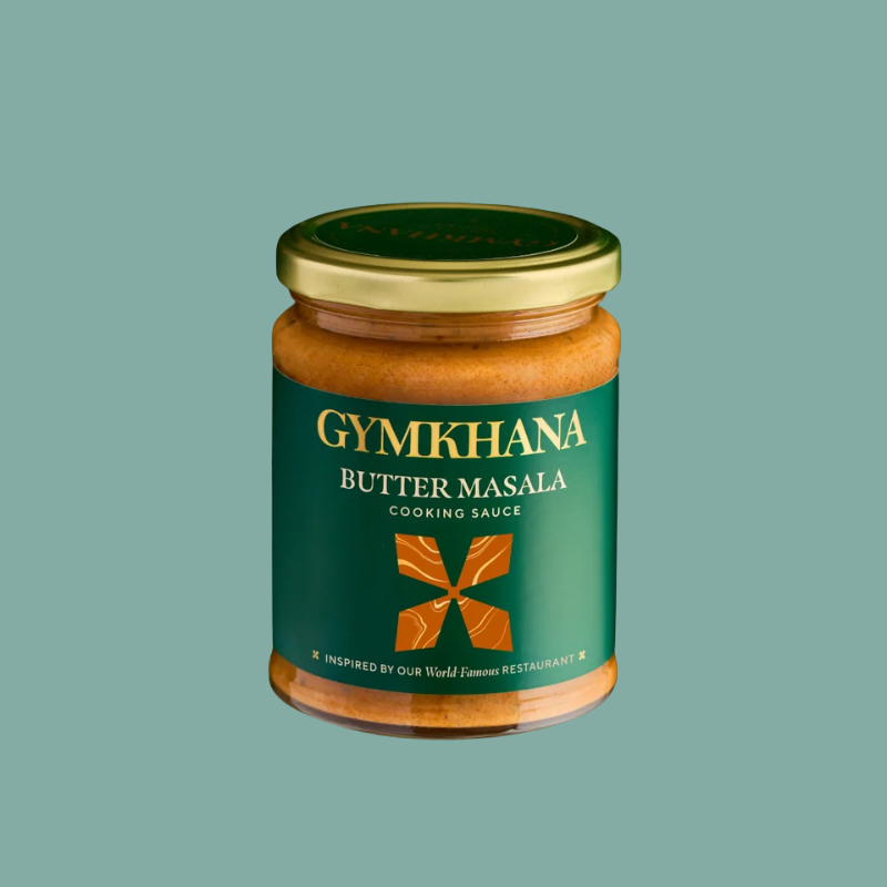 A glass jar of Gymkhana Butter Masala Cooking Sauce with a gold lid and a dark green label, centered on a solid sage green background.