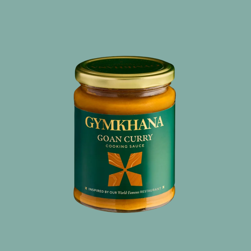 A glass jar of Gymkhana Goan Curry Cooking Sauce with a gold lid and a dark green label, centered on a solid sage green background.
