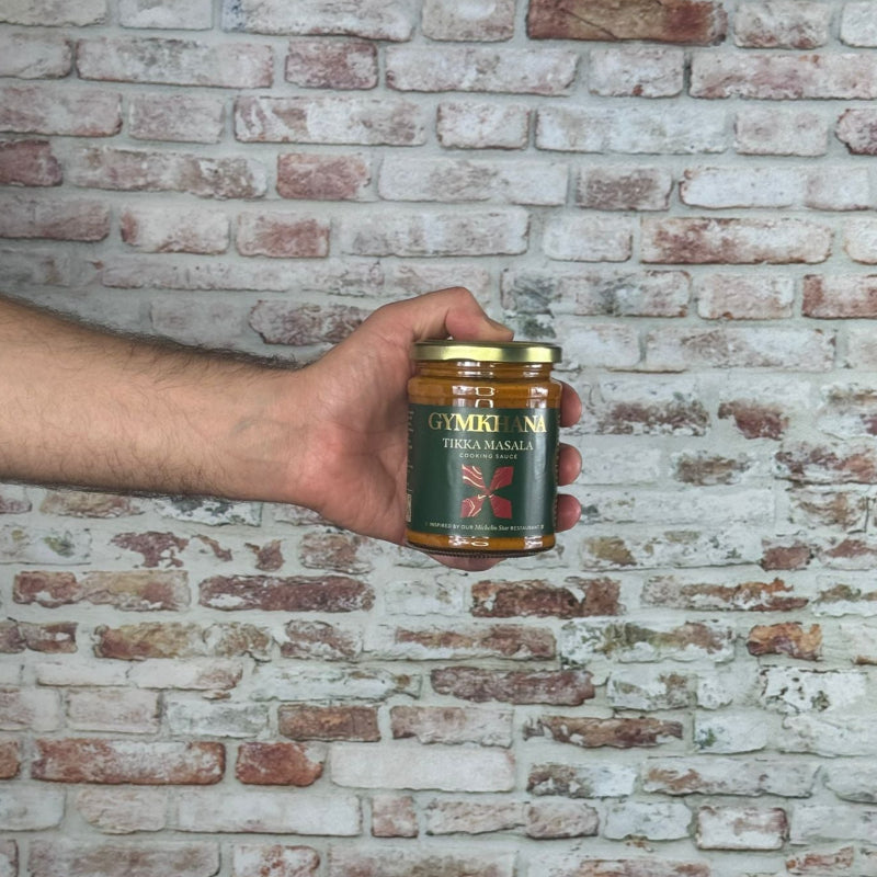 Hand holding a can of Gymkhana - Tikka Masala Sauce against a brick wall