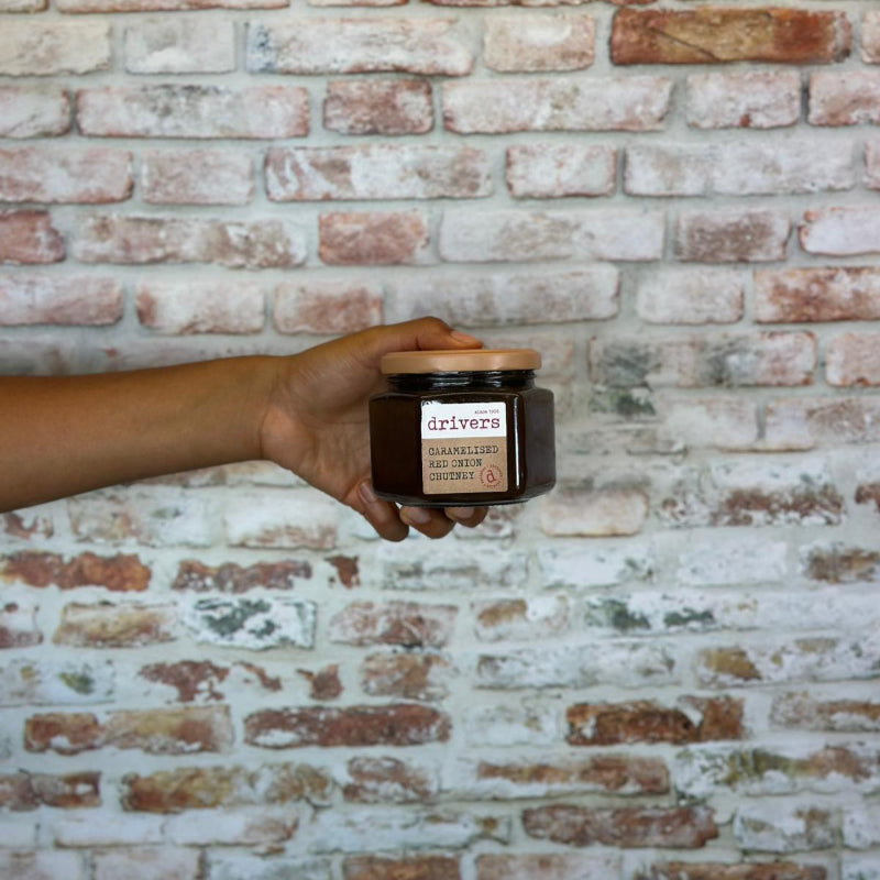 Hand holding a jar of Caramelised red onion chutney against a brick wall.