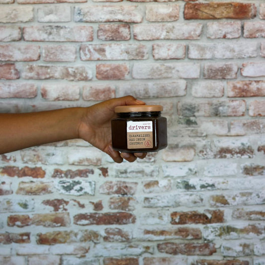 Hand holding a jar of Caramelised red onion chutney against a brick wall.