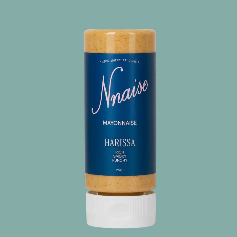 A squeeze bottle of Nnaise Harissa Mayonnaise, featuring a dark blue label with white text and a white cap, filled with orange-brown harissa paste, set against a solid teal background.