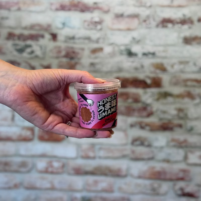 Hand holding a container of Honest Umami Chilli in front of a brick wall