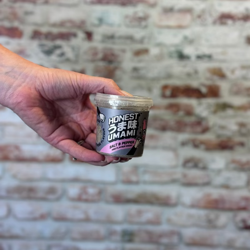 Hand holding a container of 'Honest Umami' against a brick wall background