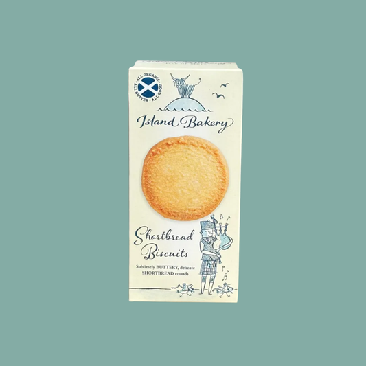 A boxed pack of Island Bakery Shortbread Biscuits on a solid teal background. The white box features blue illustrations of a cat on a roof and a bagpiper, with a photo of a round golden biscuit in the center.
