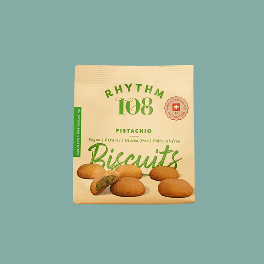 A package of Rhythm 108 Pistachio Biscuits on a solid teal background. The packaging is a light beige stand-up pouch with green text and illustrations of pistachio-filled biscuits.