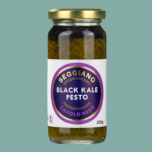 A glass jar of Seggiano Black Kale Pesto with a white and purple label and a gold metal lid, set against a solid teal background.