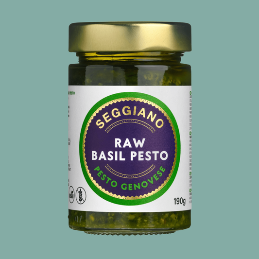 A glass jar of Seggiano Raw Basil Pesto with a gold metal lid and a white label featuring a purple and green circular logo, set against a solid teal background.