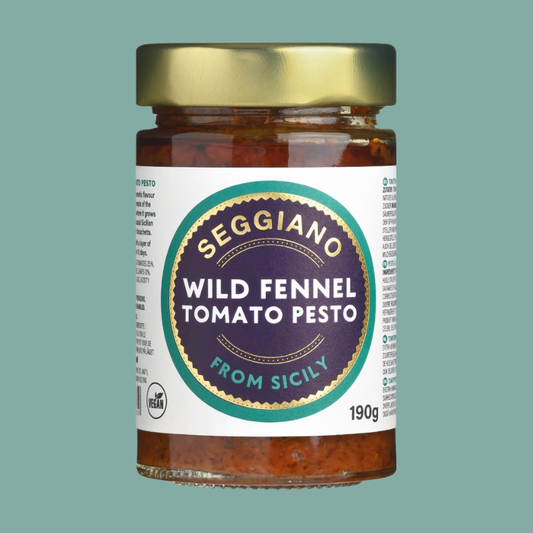A glass jar of Seggiano Wild Fennel Tomato Pesto from Sicily, featuring a white label with a prominent dark purple circular logo and a metallic gold lid, set against a solid sage green background.