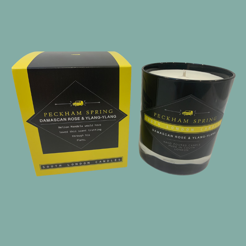 A black glass candle jar with white wax next to its yellow and black retail box. The label reads 'PECKHAM SPRING DAMASCAN ROSE & YLANG-YLANG' and 'SOUTH LONDON Candles'.