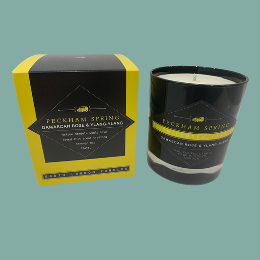 A black glass candle jar with white wax next to its yellow and black retail box. The label reads 'PECKHAM SPRING DAMASCAN ROSE & YLANG-YLANG' and 'SOUTH LONDON Candles'.