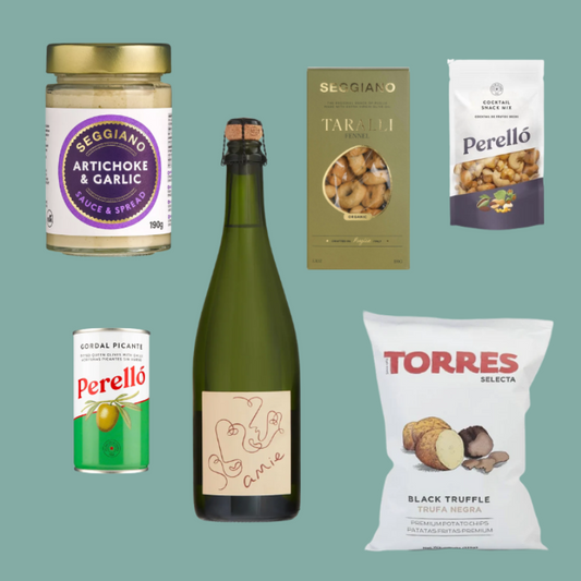 A curated collection of gourmet food products arranged on a solid sage green background, including a jar of Seggiano Artichoke & Garlic Sauce & Spreads, a bottle of Valtori wine, boxes of Seggiano Taralli and Perello pasta, a can of Perello salami, and a bag of Torres Selecta Black Truffle chips.