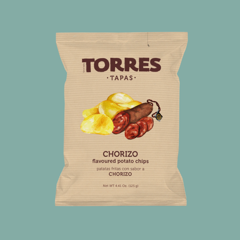 A single bag of Torres Tapas Chorizo flavoured potato chips centered on a solid muted teal background. The bag is a warm beige kraft-paper style with dark brown text and an illustration of yellow potato chips and chorizo sausage.