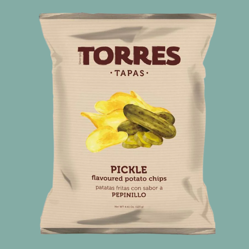 A bag of Torres Tapas Pickle flavored potato chips on a solid teal background. The packaging is a textured off-white color with dark red branding and an illustration of yellow chips and green pickled cucumbers.