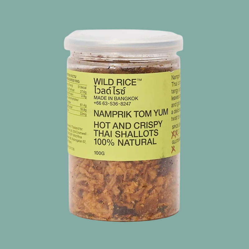 A 100g plastic jar of Wild Rice Namprik Tom Yum hot and crispy Thai shallots, featuring a yellow label and white lid, set against a solid teal background.