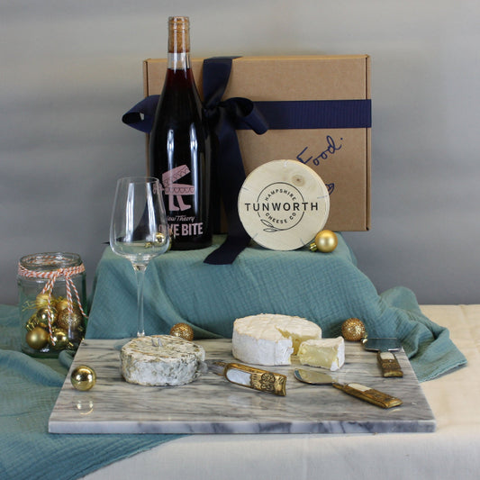 Wine bottle, cheese, and wine glass on a table with a branded box in the background.