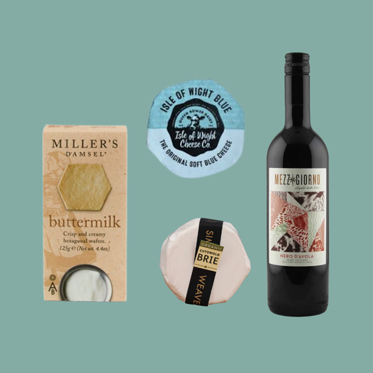 A curated food gift set on a solid teal background, featuring a box of Miller's Damsel buttermilk, a round blue sticker from Isle of Wight Blue Cheese Co., a wrapped Brie cheese wheel, and a bottle of Mezzogiorno Rosso wine.